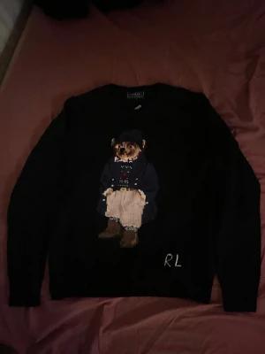 Mörkblå stickad tröja med björn från Ralph Lauren - Mörkblå stickad tröja från Ralph Lauren med ikonisk björn på framsidan, klädd i blå kappa, beige byxor och bruna boots. RL-broderi nertill. Perfekt för dig som gillar klassisk och lekfull stil.