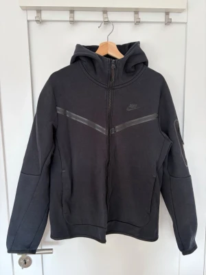 Svart Nike hoodie med dragkedja - Snygg svart hoodie från Nike med hel dragkedja framtill och huva. Hoodien har diskreta reflexdetaljer över bröstet och två fickor på sidorna. Tillverkad i mjukt och bekvämt material, perfekt för en sportig och avslappnad stil. Notera att den är lite sliten på ena armen.