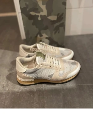 Valentino Rockrunners Vita - Snygga sneakers från Valentino i beige och vit camo-design. Ovandelen är i mesh och mocka med vita detaljer och gummisula med nitar på hälen. Perfekta för dig som gillar streetwear och exklusiva märken. Dessa skor är äkta och nypris är runt 5500kr. Dom kommer utan box och därför är priset lägre. Skriv vid minsta lilla fundering. 