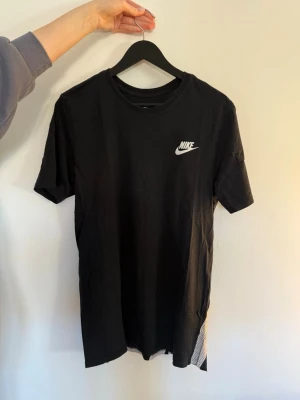 Svart Nike t-shirt med logga - Snygg svart t-shirt från Nike med vit logga på bröstet och stor Nike-text i vitt nedtill på ryggen. Klassisk passform och rund halsringning. Tillverkad i mjuk bomull som känns skön mot huden. Perfekt till streetwear eller sportig stil.