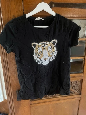 Svart t-shirt med tiger  - Cool svart t-shirt med ett stort tigerhuvud i vitt, guld och svart på bröstet. T-shirten har rund hals och korta ärmar. Perfekt för dig som vill sticka ut med ett djurmotiv och gillar streetstyle.