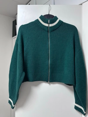 Grön cropped stickad ziptröja H&M - Mörkgrön cropped stickad tröja från H&M med dragkedja framtill. Tröjan har ribbad hög krage och muddar med vita ränder. Perfekt för en trendig och avslappnad look. Materialet är mjukt och ger en skön känsla.