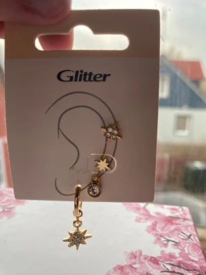 Guldiga örhängen med stjärndetaljer Glitter - Säljer ett par guldiga örhängen från Glitter med stjärnformade hängen och gnistrande stenar. Ena örhänget har en liten stjärna med kristall, det andra har en större stjärna som hänger ner. Perfekt för dig som gillar unika detaljer och glittriga accessoarer.