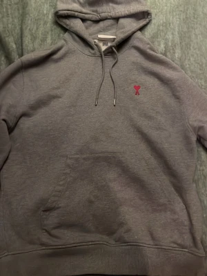 Ami Paris hoodie - Säljer en grå hoodie från AMI Paris med den ikoniska röda hjärtloggan broderad på bröstet. Hoodien har huva med dragsko, magficka och ribbade muddar. Tillverkad i mjuk bomullsblandning som är riktigt skön att ha på sig. Ger vintage känsla över hoodien. Skriv för mått