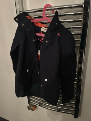 Mörkblå vindjacka från 2117 of Sweden - Snygg mörkblå vindjacka från 2117 of Sweden med dragkedja, tryckknappar och två stora fickor framtill. Jackan har huva och orange detaljer på insidan samt logga på bröstet. Perfekt för blåsiga dagar och enkel att matcha med olika outfits.