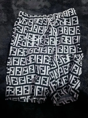 Fendi shorts - Säljer ett par svarta och vita shorts från Fendi med ikoniskt FF-mönster över hela plagget. Shortsen har elastisk midja med vit snörning och är tillverkade i ett lätt och bekvämt material, perfekta för sommardagar.