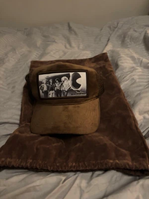 Brun keps med Maggiore-tryck - Snygg brun keps med svartvit patch framtill som visar ett retroinspirerat foto av ett band och texten 'MAGGIORE'. Kepsen har böjd skärm och mesh på sidorna för extra ventilation. Perfekt accessoar för en cool och avslappnad stil.