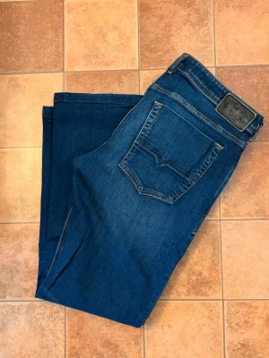 Diesel Zatiny Bootcut Jeans - W36 L34 - Säljer ett par klassiska Diesel Zatiny i en snygg mörkblå tvätt med lätta slitningar. Modellen är en populär Micro-Bootcut (svag utställd nertill) som ger en riktigt snygg siluett. Detaljer: • Modell: Zatiny • Storlek: W36 / L34 • Passform: Bootcut / Regular fit • Tvätt: 082AY (Mörkblå med subtil stretch) • Material: 98% Bomull, 2% Elastan (ger bra komfort) • Skick: Mycket gott skick, väl omhändertagna. • Autenticitet: Givetvis äkta – Certilogo-tagg finns i byxan (se bild). 