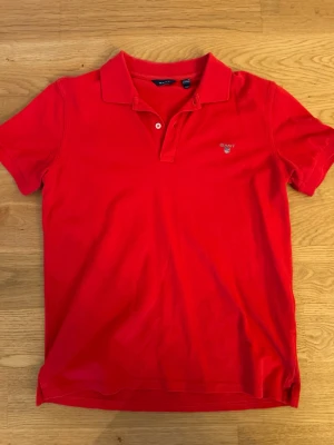 Pikétröja - Säljer en klassisk röd pikétröja från GANT med krage och två knappar framtill. Tröjan har broderad logotyp på bröstet och är tillverkad i bomull. Perfekt för dig som gillar stilrena och sportiga plagg.