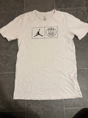 Vit PSG x Jordan T-shirt - Säljer en vit T-shirt från Jordan i samarbete med Paris Saint-Germain. T-shirten har en klassisk passform, rund halsringning och tryck med Jordan-loggan samt PSG-emblem på bröstet. Perfekt för dig som gillar sportig streetwear.