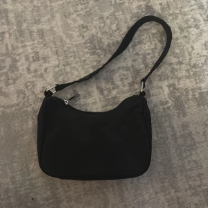 Svart axelväska från Gina Tricot - Stilren svart handväska från Gina Tricot med  tygmaterial och silverfärgade detaljer. Väskan har en böjd form, dragkedja upptill och bred axelrem som gör den enkel att bära. Perfekt för att hålla dina essentials nära.