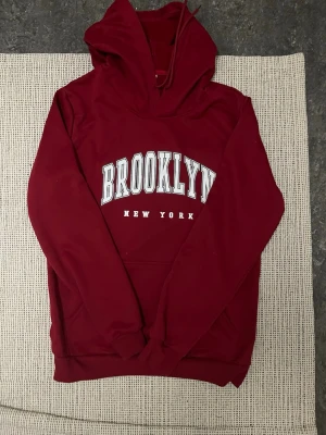 Vinröd hoodie med Brooklyn-tryck - Snygg vinröd hoodie med stor vit text 'BROOKLYN' och mindre 'NEW YORK' på bröstet. Klassisk känguruficka framtill och justerbar huva med dragsko. Perfekt för en avslappnad streetwear-look. Mjuk bomullsblandning för extra komfort.