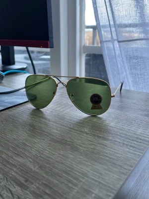 Ray-Ban aviator solglasögon guld/grön - Snygga Ray-Ban aviator solglasögon med tunna guldfärgade metallbågar och klassiska gröna glas. Ikonisk pilotform som ger en tidlös look. Perfekta för soliga dagar och ger UV-skydd. Ray-Ban-logga syns på linsen.