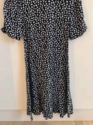 Blommig mörkblå klänning med kort ärm - Supersöt mörkblå klänning med vitt blommönster och korta ärmar. Klänningen har en lätt utställd passform och volangkant nertill. Perfekt för dig som gillar en feminin och avslappnad stil. Materialet är skönt och luftigt