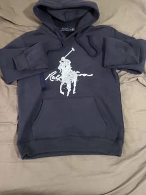 Mörkblå hoodie från Polo Ralph Lauren - Snygg mörkblå hoodie från Polo Ralph Lauren med stor vit logga och polospelare på bröstet. Klassisk känguruficka framtill, justerbar dragsko i huvan och ribbade muddar. Perfekt för en avslappnad och trendig stil.