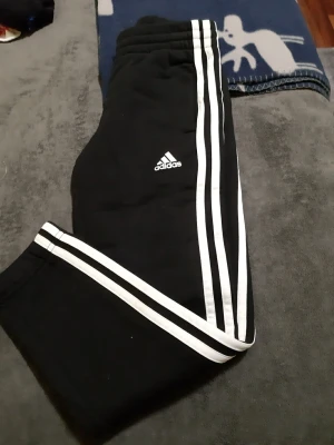 Helt nya, aldrig används. Svarta Adidas mjukisbyxor med vita ränder - Svarta mjukisbyxor från Adidas med klassiska vita ränder längs sidorna och Adidas-logga på låret. Byxorna har resår i midjan och är tillverkade i mjukt material för maximal komfort. Perfekta för en avslappnad och sportig stil. Helt nya.