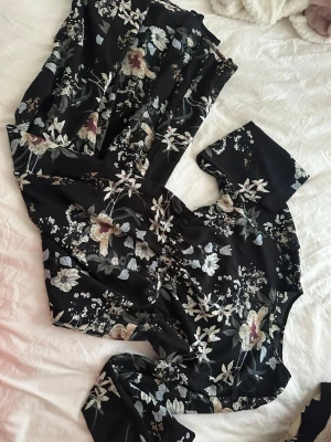 Svart blommig långklänning - Svart långklänning med blommigt mönster i beige, vitt och ljuslila. Klänningen har lång ärm och markerad midja för en snygg siluett. Perfekt för dig som gillar romantiska och feminina detaljer.