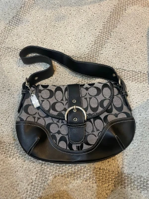 Coach axelväska med monogram - Snygg axelväska från Coach i svart och grått med klassiskt monogrammönster. Väskan har detaljer i svart läder, silverfärgat spänne och bred axelrem. Perfekt för dig som gillar stilrena accessoarer med en touch av lyx. Superfin❤️