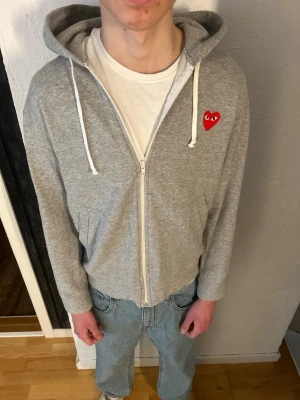 Grå hoodie Comme des Garçons Play - Säljer en grå zip-hoodie från Comme des Garçons Play med röda hjärtapplikationer och logga på ryggen samt ett litet hjärta på bröstet. Hoodien har dragsko i huvan och är tillverkad i mjukt bomullsmaterial. Perfekt för en avslappnad och trendig look.