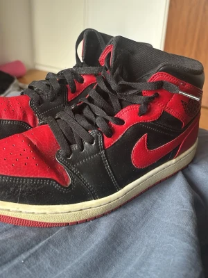 Air Jordan 1 Mid svart/röd - Säljer ett par Air Jordan 1 Mid i klassisk svart och röd färgkombination med vita sulor. Perfekta för dig som gillar streetwear och sneakerskultur.