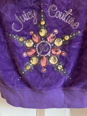 Lila Juicy Couture hoodie med diamantstenar - Säljer en lila hoodie från Juicy Couture i mjuk velour. Baksidan har ett stort stjärnmönster dekorerat med glittriga stenar i rosa och gult samt broderad text. Hoodien har huva och långa ärmar, perfekt för dig som gillar vintage-plagg och bling.