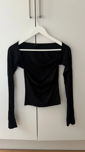 Svart långärmad topp med drapering - Trendig svart topp med lång ärm och snygg drapering framtill. Modellen är figurnära och har en bred, något djupare urringning. Perfekt att styla med jeans eller kjol för en cool look.