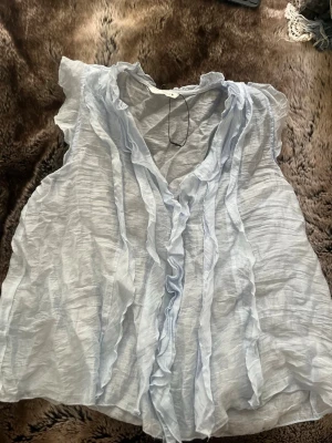 Ljusblå volangblus i tunn bomull - Säljer en superfin ljusblå blus i tunn, luftig bomull. Blusen är ärmlös och har snygga volangdetaljer framtill samt en v-ringad hals. Perfekt för varma dagar och ger en romantisk vibe till din outfit.