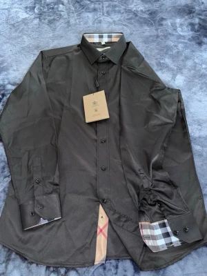 Svart skjorta från Burberry - Snygg svart skjorta från Burberry med klassiskt rutigt mönster på insidan av kragen och manschetterna. Skjortan har knappar framtill och en diskret broderad logga på bröstet. Tillverkad i ett mjukt och glansigt material som känns lyxigt.