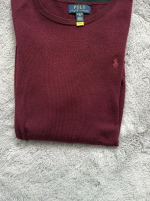Vinröd stickad tröja Polo Ralph Lauren - Snygg vinröd stickad tröja från Polo Ralph Lauren med rund halsringning och diskret broderad logga på bröstet. Tröjan har långa ärmar och ribbade muddar vid ärmslut och nederkant. Perfekt för dig som gillar klassisk och stilren design. Storlek XL 18-20 för ungdom, sitter som en XS/S tröja