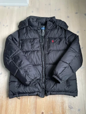 Svart pufferjacka från Polo Ralph Lauren - Säljer en svart pufferjacka från Polo Ralph Lauren med huva och dragkedja. Jackan har det klassiska röda Polo-logotypen broderad på bröstet och är tillverkad i ett glansigt, slitstarkt material. Perfekt för kalla dagar och riktigt snygg till streetwear-stilen. Använd i ca 2 månader men fortfarande i utmärkt skick. Storlek medium för vuxna. Jag är 184 och den sitter perfekt