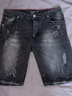 Svarta jeansshorts från  Philipp Plein  - Säljer ett par svarta jeansshorts från Philipp Plein med slitningar, färgstänk och råa kanter. Shortsen har fem fickor och ett gult PP-märke på bakfickan. Perfekta för dig som gillar en edgy streetstil.
