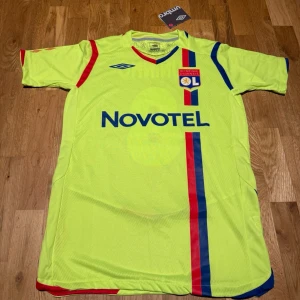 Lyon away kit 09/10 X Juninho - Säljer en officiell Olympique Lyonnais matchtröja från Umbro med Juninho och nummer 8 på ryggen. Tröjan är gul med blå och röda detaljer, klubbmärke på bröstet och Novotel-tryck framtill. Perfekt för fotbollsfans som vill sticka ut på planen eller läktaren.