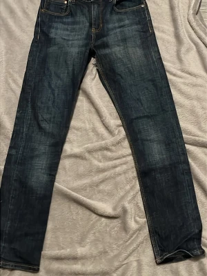 Mörkblå jeans från Drain - Snygga mörkblå jeans från Drain med raka ben och klassiska fem fickor. Jeansen har kontrastsömmar i gult och metallknapp med märkets logga. Perfekta för dig som gillar en enkel och stilren look. De har även ett litet hål vid sidan av låret 