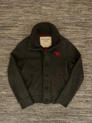 Mörkgrå kofta från Abercrombie & Fitch - Mörkgrå kofta från Abercrombie & Fitch i storlek S med hög krage, knäppning framtill och två fickor. Koftan har den klassiska älg loggan på bröstet och ribbade muddar. Perfekt för lager på lager och en chill look. Skriv vid minsta fråga eller fundering! Priset kan alltid diskuteras!