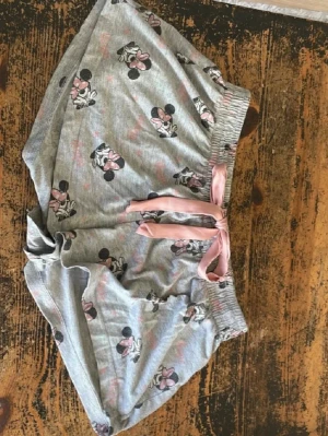 Grå Minnie Mouse pyjamasshorts - Supersöta grå pyjamasshorts från Disney med Minnie Mouse-motiv och rosa detaljer. Shortsen har ett elastiskt midjeband och en rosa rosett framtill. Perfekta för mysiga hemmakvällar eller sommarens varma nätter. Tillverkade i mjukt bomullsmaterial.