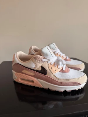 Nike Air Max 90 rosa och beige sneakers - Nike Air Max 90 sneakers i vitt, beige och olika nyanser av rosa med detaljer i brunt. Ovandelen är i mesh och läder, med klassisk synlig Air-enhet i sulan. Swoosh-logga på sidan och gummisula med bra grepp. Perfekta för dig som gillar stilrena men färgglada sneakers.