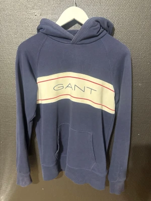 Blå hoodie från GANT  - Snygg blå hoodie från GANT med klassisk logga på bröstet och kontrasterande vit panel med röda linjer. Hoodien har huva, raglanärmar och en stor magficka. Perfekt för dig som gillar stilrena och sportiga plagg.