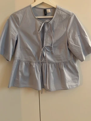 Ljusblå blus med knyt från H&M Divided - Supersöt ljusblå blus från H&M Divided med knytband framtill och volang nedtill. Blusen har korta ärmar och en avslappnad passform, perfekt för en trendig look. Tillverkad i ett lätt och mjukt tyg (100% bomull) som känns skönt mot huden.