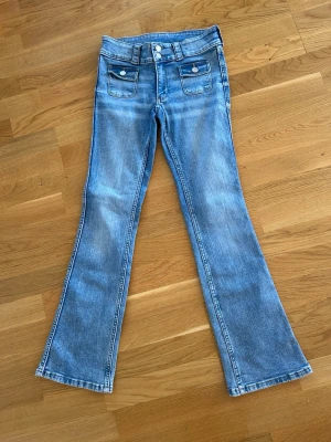 Ljusblå bootcut jeans med fickor - Säljer ett par ljusblå bootcut jeans med dubbla framfickor med lock och knappar. Jeansen har en klassisk femficksdesign, låg midja och utsvängda ben. Perfekta för dig som gillar retrostil och vill ha ett par snygga jeans i denim.