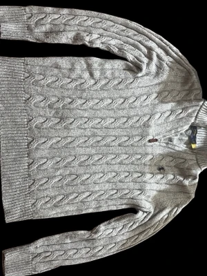 Quarter zip Ralp lauren  - Snygg grå kabelstickad tröja från Polo Ralph Lauren med klassisk rund hals och ribbade muddar. Tröjan har det ikoniska lilla broderade logotypen på bröstet och är tillverkad i mjukt garn som känns skönt mot huden.