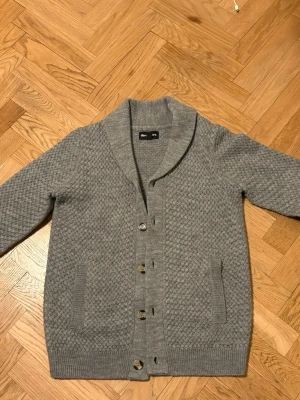 Grå kofta från Threadbare - Snygg grå kofta från Threadbare med sjalkrage och grovstickat mönster. Koftan har knäppning med fem knappar framtill och två fickor. Perfekt för lager på lager och chill stil. Nyrpis 579. PRIS KAN DISKUTERAS SKRIV PRIVAT FÖR MER FRÅGOR