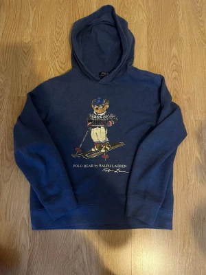 Blå hoodie Polo Bear Ralph Lauren - Mörkblå hoodie från Ralph Lauren med Polo Bear-tryck på bröstet där björnen åker skidor. Klassisk huva och ribbade muddar. Mjuk bomullskänsla och sportig look, perfekt för dig som gillar streetwear och ikoniska prints. Storlek 164-170! Skick 9/10 bra skick! Skriv till mig vid intresse eller frågor!
