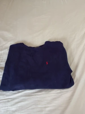 Mörkblå sweatshirt från Polo Ralph Lauren - Klassisk mörkblå sweatshirt från Polo Ralph Lauren med rund halsringning och den ikoniska röda logotypen broderad på bröstet. Tröjan har långa ärmar och ribbade muddar vid ärmslut och nederkant. Perfekt för en avslappnad och stilren look.