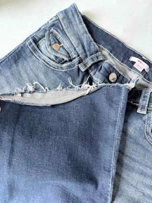 Blå jeans från Gina Tricot - Snygga blå jeans från Gina Tricot med klassisk femficksmodell och råa kanter vid linningen. Jeansen har en mörk tvätt och silverfärgade knappar