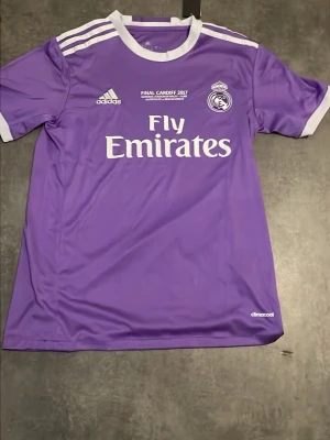 Lila Real Madrid tröja Adidas - Säljer en lila Real Madrid matchtröja från Adidas med vita detaljer och tryck. Tröjan har klubbmärke, Adidas-logga och Fly Emirates sponsortryck på bröstet samt texten 'Final Cardiff 2017'. Tillverkad i lätt och ventilerande material.
