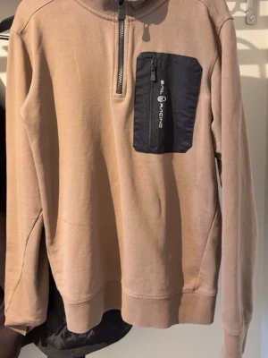 Beige Sail Racing hoodie med zip - Säljer en beige hoodie från Sail Racing med halv dragkedja framtill och en svart bröstficka med logga och zip. Hoodien har ribbade muddar och är gjord i mjukt sweatshirtmaterial. Perfekt för en avslappnad och sportig stil.