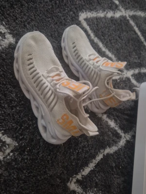 Vita stickade sneakers med text - Snygga vita sneakers med stickad ovandel och unik sula i vågig design. Skorna har orange textdetaljer på sidan och tungan. Lätta och luftiga med snörning och högt skaft för extra stöd.