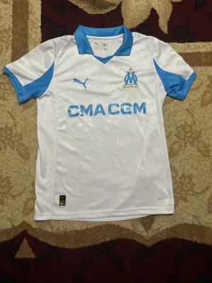 Marseille matchtröja Puma vit/blå - Snygg vit matchtröja från Olympique de Marseille med ljusblå detaljer och Puma-logga. Tröjan har klubbmärke och sponsortryck på bröstet samt diskreta klubbmotiv i tyget. Perfekt för fotbollsträning eller att bära till vardags.