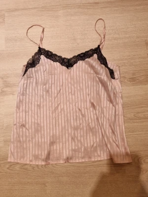 Ljusrosa randig linnetopp med spets - Säljer en ljusrosa linnetopp med tunna axelband och svarta spetsdetaljer vid urringningen. Toppen har ett subtilt randigt mönster och är gjord i ett glansigt material som känns lyxigt och mjukt. 