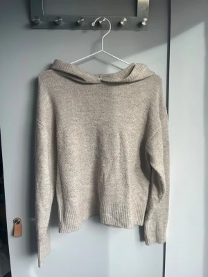 Beige stickad hoodie - Mysig beige stickad hoodie med avslappnad passform och ribbade muddar vid ärmslut och nederkant. Perfekt för lager på lager och chill dagar. Huvan är bred och ger en skön look. Materialet känns mjukt och värmande.